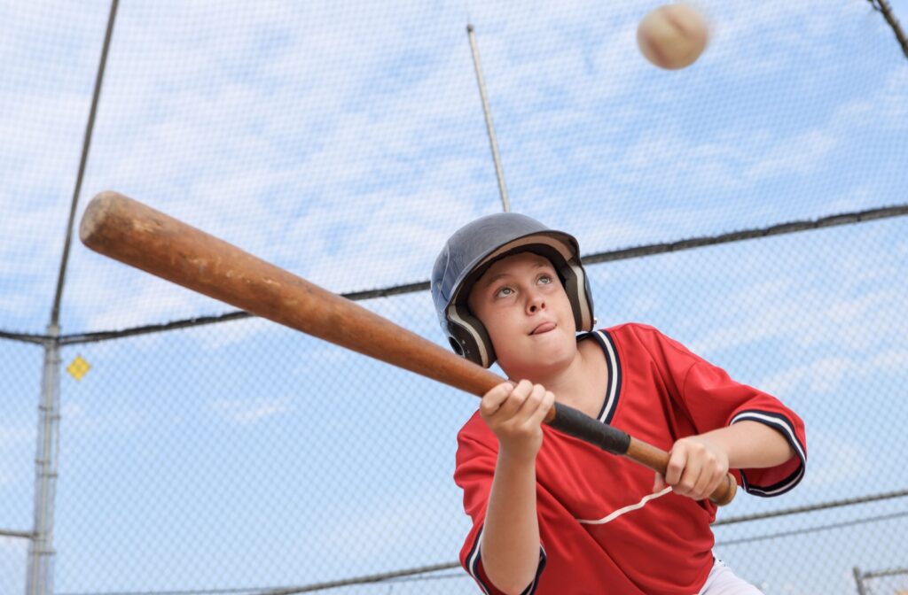 A young boy in red sticks his tongue out in concentration as he attempts to hit an incoming baseball with a wooden bat.