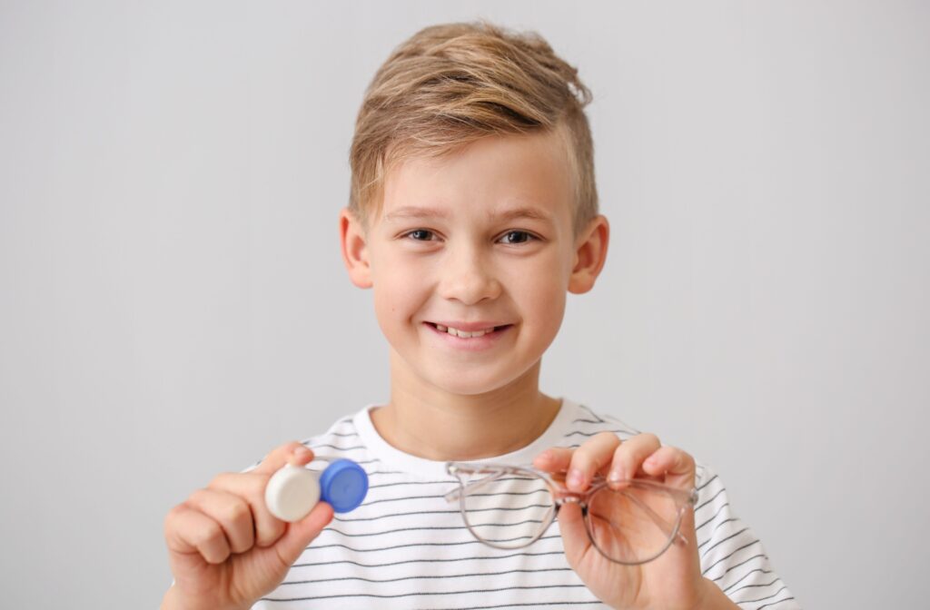 A child smiles into the camera, holding up a pair of glasses in one hand and a contact lens case in the other.