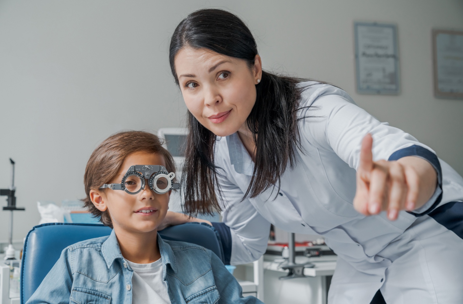 A child sits in an optometrist's chair, looking through a phoropter. The optometrist stands beside the child, pointing at an eye chart outside the picture.