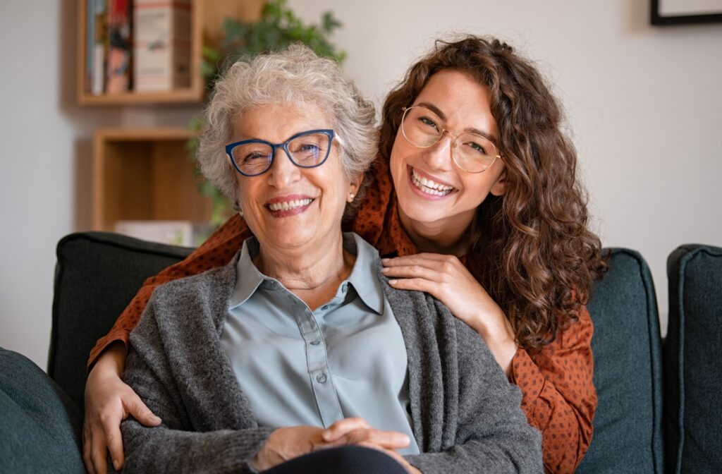 A young adult with glasses cheerfully embraces an elderly loved one, who is also wearing glasses.